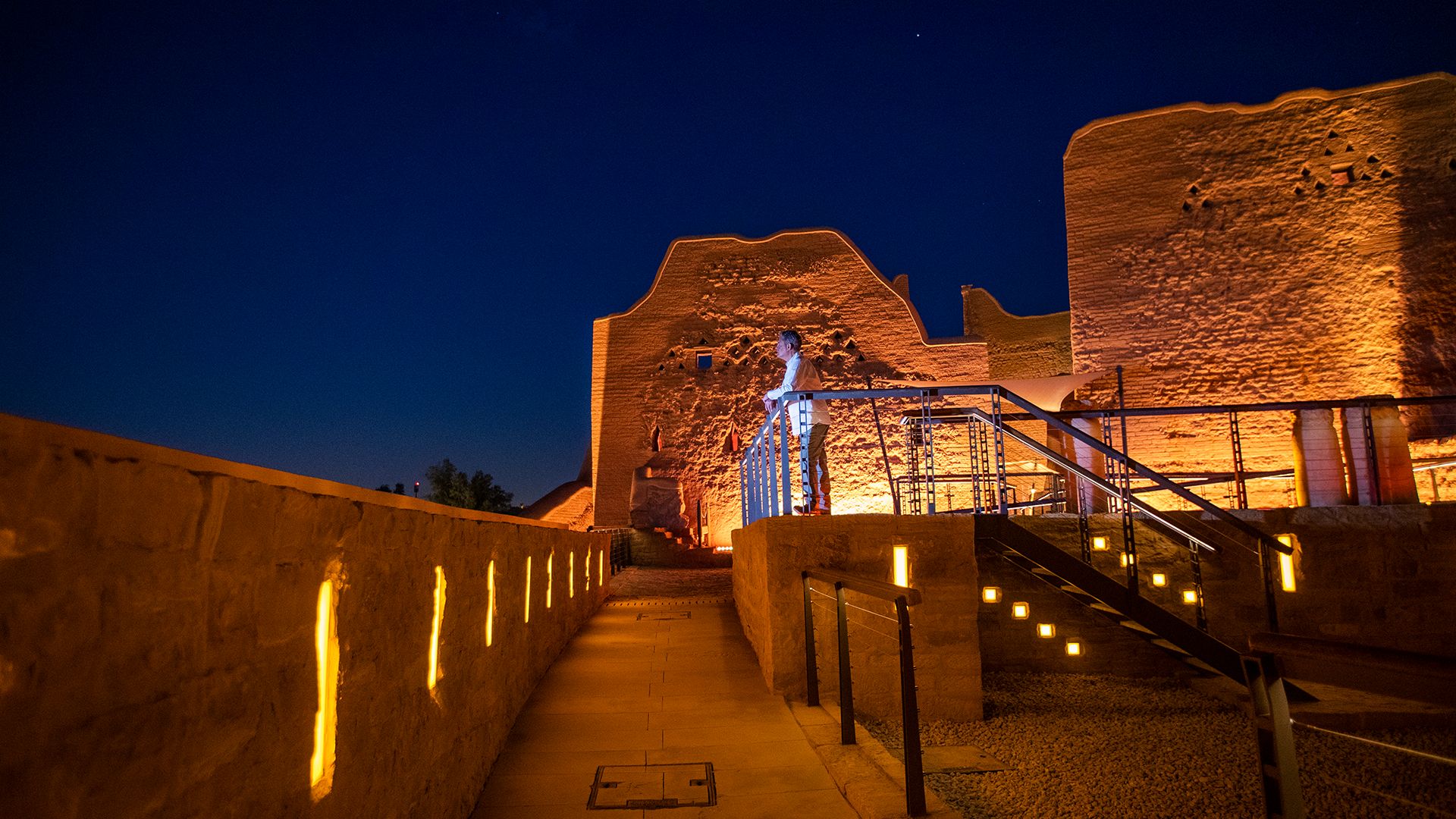 Night view of Diriyah’s mudbrick heritage walls and a lit walkway, with a person standing near the historic structure.